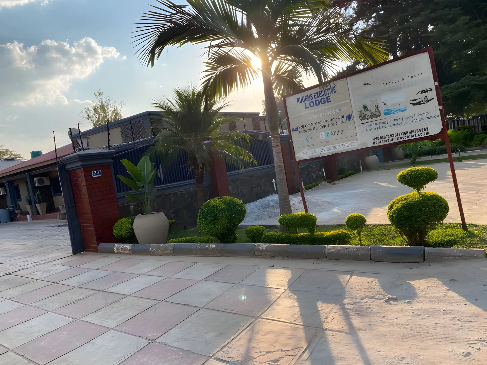 Rugems Executive Lodge entrance at golden hour — palm tree, gate and driveway